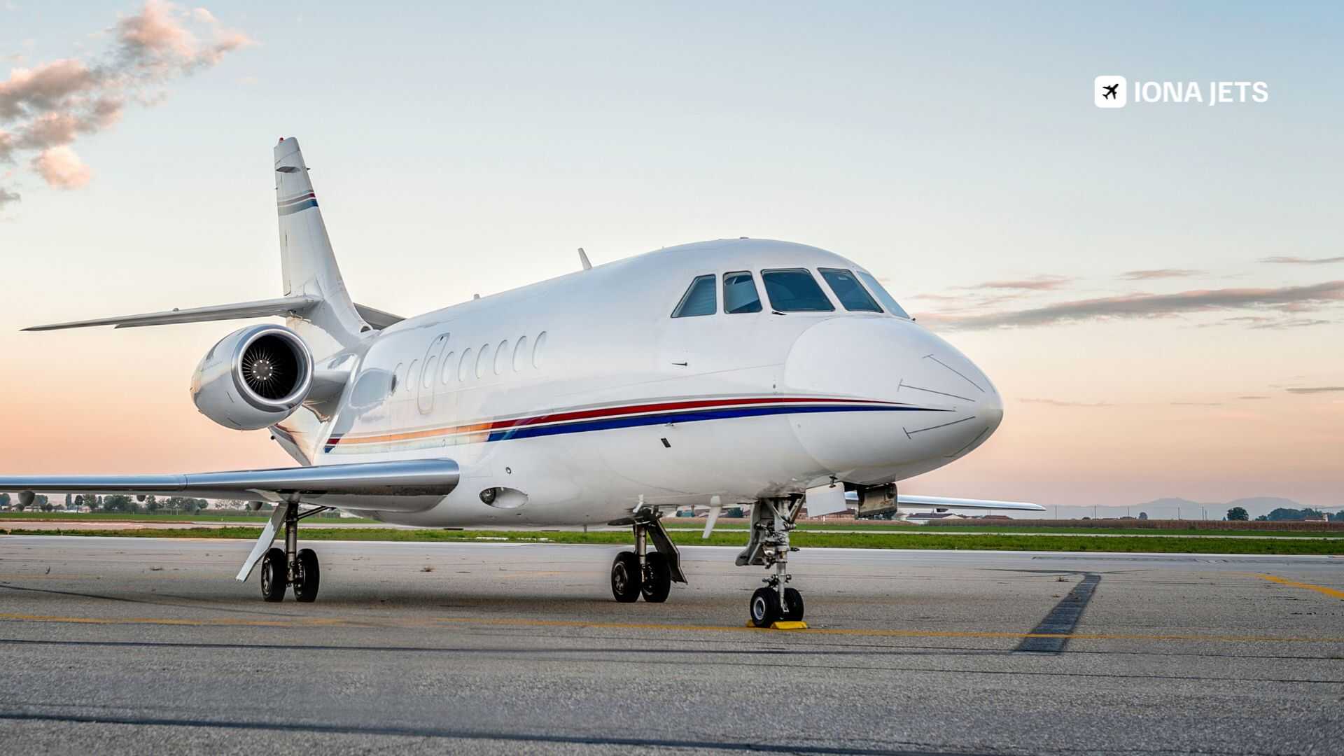 Side-front view of a white business jet on the tarmac at sunset, showing engines and landing gear, with open landscape, soft sky and IONA JETS branding in the corner.